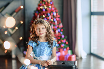 girl with dark hair standing on a box with gifts. Christmas tree in the background. smiles