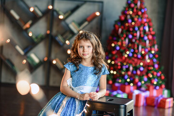 girl with dark hair standing on a box with gifts. Christmas tree in the background. smiles