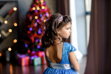 girl with dark hair standing on a box with gifts. Christmas tree in the background. smiles