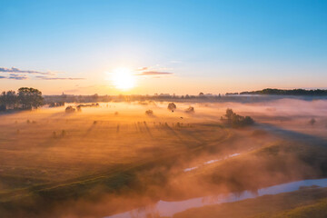 Morning fog in the meadows and at the mouth of the river during sunrise in the countryside, aerial view height of the landscape on the horizon, a forest and a village with flying birds.