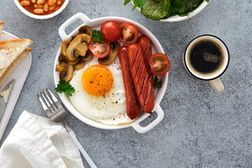 Traditional full English breakfast with fried eggs, sausages, beans, mushrooms, grilled tomatoes on grey background.