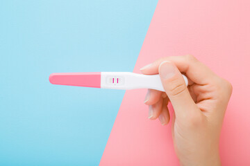 Young woman hand holding pregnancy test with two stripes on light pink blue table background. Pastel color. Two sides. Positive result. Closeup. Point of view shot. Top down view.