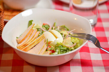 Chicken Caesar salad served in a white bowl. Traditional salad with chicken meat, eggs, tomatoes. Dinner in restaurant.
