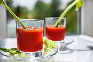 Two glasses of tomato juice with parsley and celery decorations, stand near the window, morning sunlight shines, shallow depth of field, selective focus. Natural drinks concept.