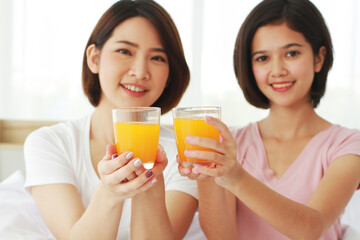 Asian young sisters lovely couple on white bed and smiling with hand holding Orange juice glass in bedroom. Holiday and healthy concept. Closed up on Orange juice glass.