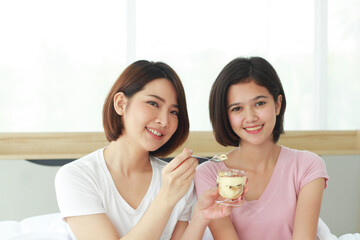 Asian young sisters lovely couple on white bed hand holding cup of cake and eating Bakery together in bedroom. Homosexual women or Lesbian in love. LGBT Asian and Holiday concept.