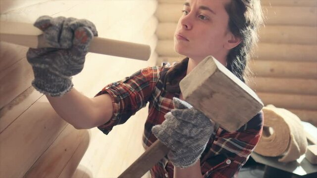 Young woman caulking and sealing wooden logs with oakum. Pretty woman works inside the wooden building and operates with hammer. Young feminist girl worker. Gender equality concept