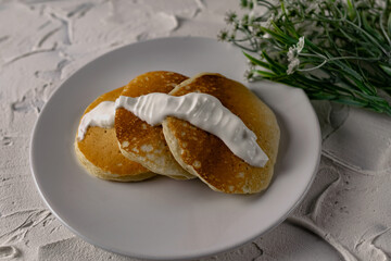 Crumpets and pancakes with sour cream on a white plate with white sauce on top. Next to a sprig of fresh light flowers and compote. Interesting textured background of white stone
