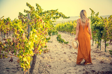 
Blonde back woman with long blonde hair and hat in hand walks through a vineyard farm