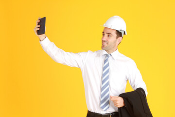 Handsome smart engineer or architecture is Wearing a white safety hat with hand holding smartphone and enjoy with Selfie by himself isolated on yellow background. Copy Space