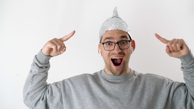 White man in a foil hat smiles and joyfully points his fingers at the tinfoil hat. 5G tower radiation protection. Irrational fear of a non-existent problem. Protective helmet to the brain from aliens.
