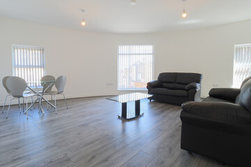 A modern British living room with two black leather sofas and a dining table on a grey wooden floor with white walls and modern blinds up at the window