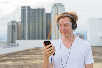 Fototapeta na wymiar Happy young man with face shield using phone and listening to music at rooftop of the building