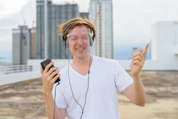 Fototapeta na wymiar Happy young handsome man with face shield dancing while listening to music at rooftop of the building