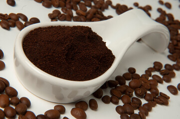 Recipiente blanco con café molido rodeado de granos de café sobre fondo blanco
