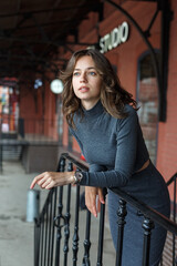 young thoughtful pretty girl in gray turtleneck, skirt with watches on arm poses in front of red building. Street style portrait photosession of attractive female, urban photoshoot of elegant model