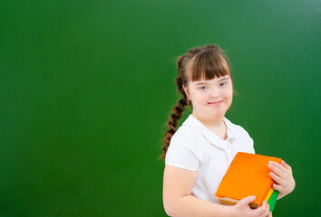 Girl with Down Syndrome standing against a green school board and holding a stack of books. Education accessible to everyone