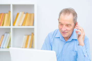 Fototapeta premium Handsome elderly man working in an office on a laptop talking on a cell phone with a smile looking at a computer screen