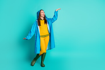 Full length photo of attractive lady toothy smile cheerful look up check rain drops arm rainy weather street walk wear casual outfit raincoat hood gumboots isolated teal color background