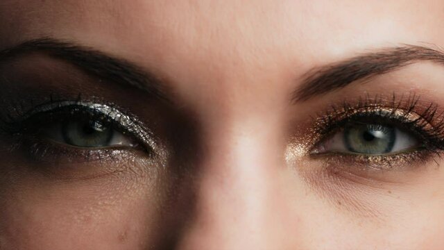 Female with beautiful makeup, glitter shadows and false lashes opening eyes. Womens green eye contracting. Extreme close up of human face in 4K video. 