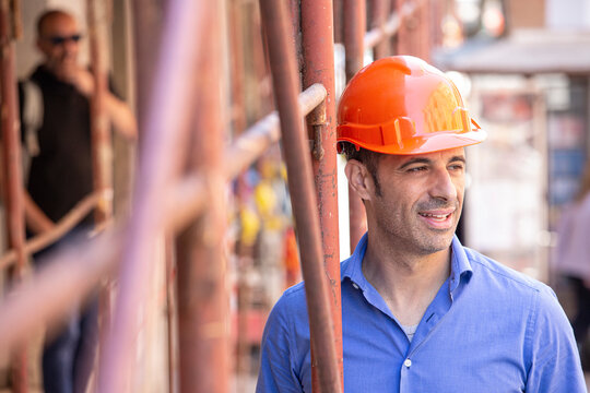 Uomo con camicia blue e casco arancione  sorride sotto il ponteggio di un cantiere in citt&agrave;