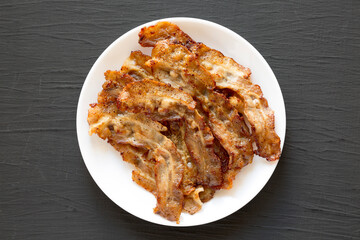Homemade Fried Bacon on a white plate on a black surface, top view. Overhead, from above, flat lay.
