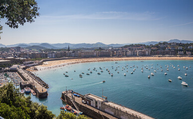 Fototapeta premium Panoramic view of a bay with a small port and a beach with people swimming, with some boats in the sea on a quiet summer day