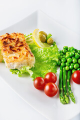 Cod fillet baked with cheese and onions with a garnish from an asparagus and green peas. On white background. High key. Selective focus.