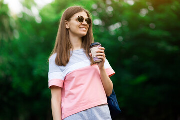 Portrait of beautiful brunette girl in sunglasses walking down the street. Keeping takeaway drink in hand. Smiling.