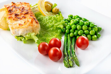 Cod fillet baked with cheese and onions with a garnish from an asparagus and green peas. On white background. High key. Selective focus.