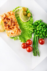 Cod fillet baked with cheese and onions with a garnish from an asparagus and green peas. On white background. High key. Selective focus.