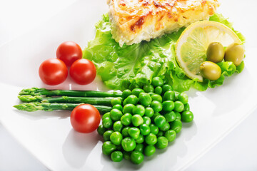 Cod fillet baked with cheese and onions with a garnish from an asparagus and green peas. On white background. High key. Selective focus.