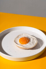 Fried egg on a white plate on a yellow background with shadows.