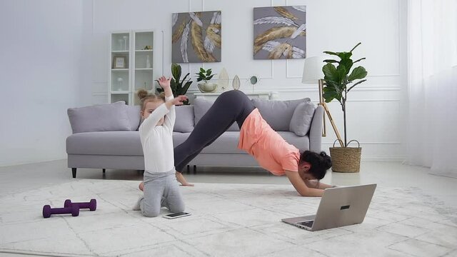 Likable athletic flexible young woman doing cat yoga pose while her smiling small daughter watching funny cartoon on phone