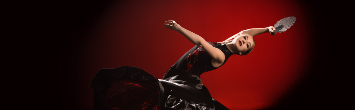 panoramic shot of young flamenco dancer in dress holding fan while dancing on red