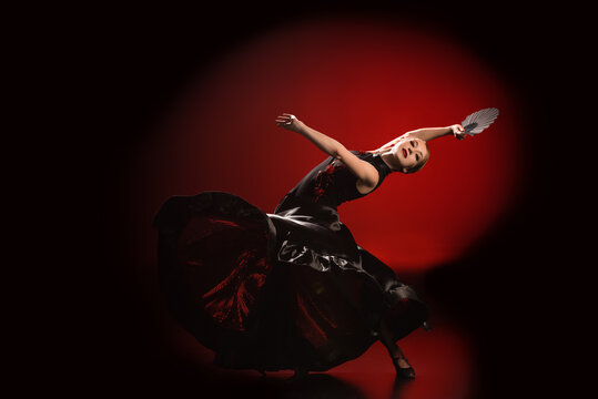 young flamenco dancer in dress holding fan while dancing on red and black
