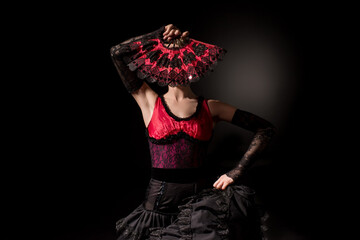 young flamenco dancer in dress covering face with fan on black