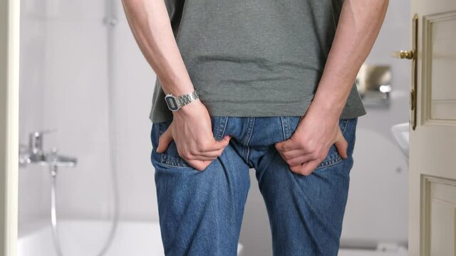 Tall man in restroom feels sudden stinging pain in anus area. Hands on buttocks, tearing jeans. Medium shot, real time