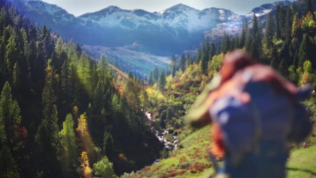 Young redhead woman taking photos on a sunny autumn day during a hike, wearing a backpack. Alpine mountains and trees with green pastureland in the background. Focus racking.