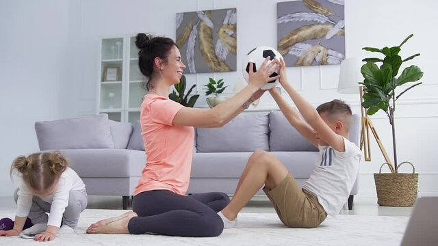 Front view of lovely smiling young woman which helping to do aps with her son using ball and her one-year nice daughter playing near them