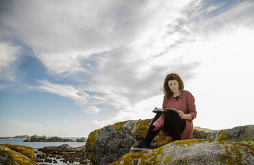 Naklejka na ściany i meble Black-haired woman reading book on the rocks by the sea