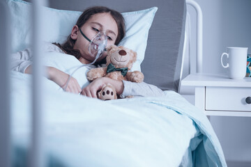 Girl suffering from pneumonia lying in a hospital bed with oxygen mask and teddy bear