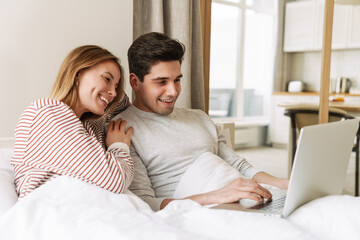 Portrait of joyful couple smiling and using laptop while lying in bed