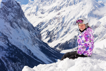 Fototapeta na wymiar Cute teen girl sit in snow on top of the mountain in ski outfit with helmet and mask turn back smiling to camera