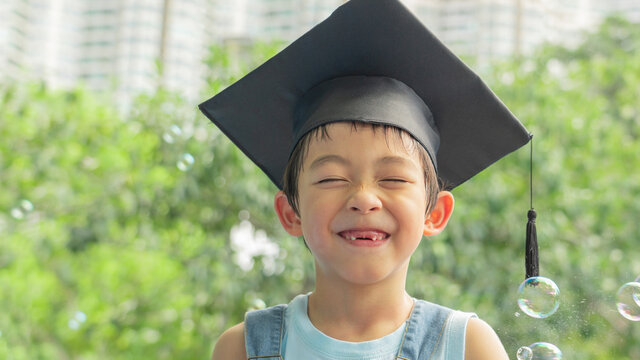 One happy and cute boy with the graduation cap/hat on is closing his eyes and smiling with fun at the park. Kids and the front milk teeth missing at outdoor play area.