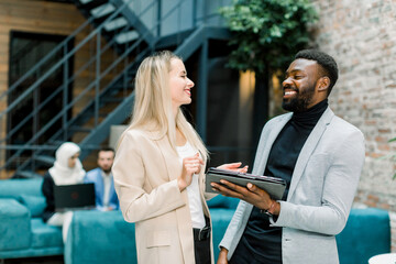 Group of happy smiling multiethnical business people, pretty blond Caucasian woman and handsome bearded African man, standing together, laughing and enjoying work, sharing their ideas.