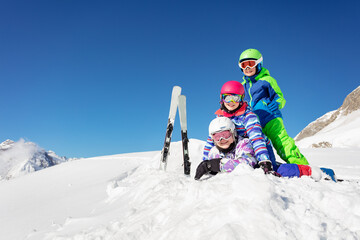 Pile of kids one on top of another lay in the snow over blue sky in the mountains with ski on background