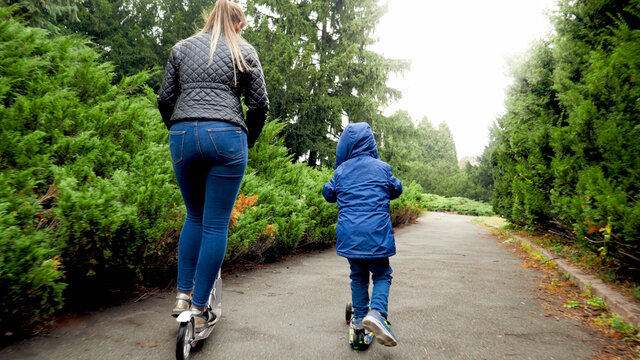 Rear view image of little boy with young mother riding on scooters on pathway at park