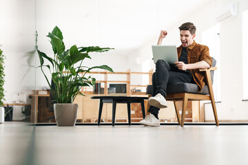 Image of delighted man making winner gesture while working with laptop