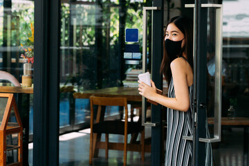 Young beautiful Asian woman wearing a surgical face mask while leaving a coffee cafe shop. Attractive female customer buying coffee and holding a disposable coffee cup. Covid-19 prevention.